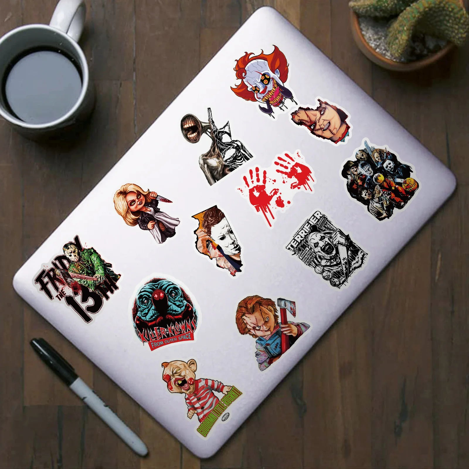 Laptop with collection of horror film stickers on a wooden surface with a cup of coffee and pen.