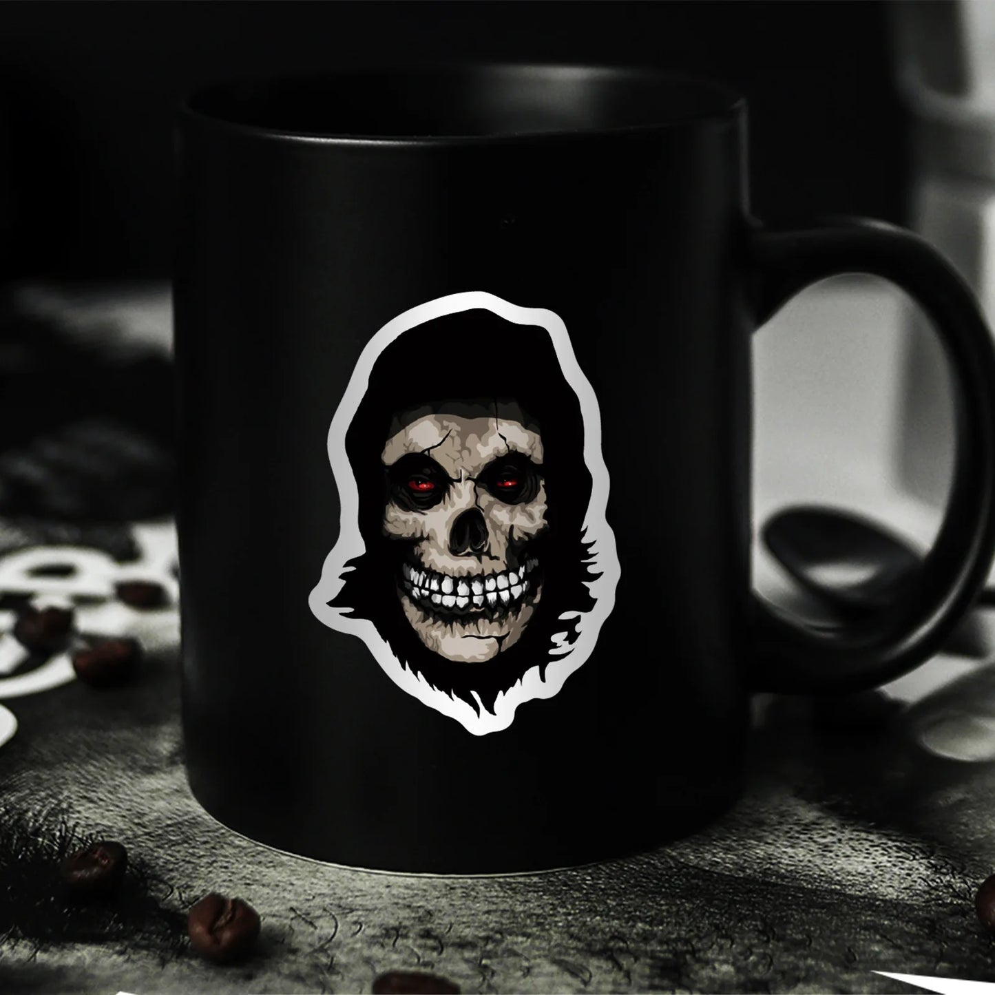 Black mug with one of the horror film sticker set stickers and one on the mug of a skull design on a dark surface with coffee beans in the background.