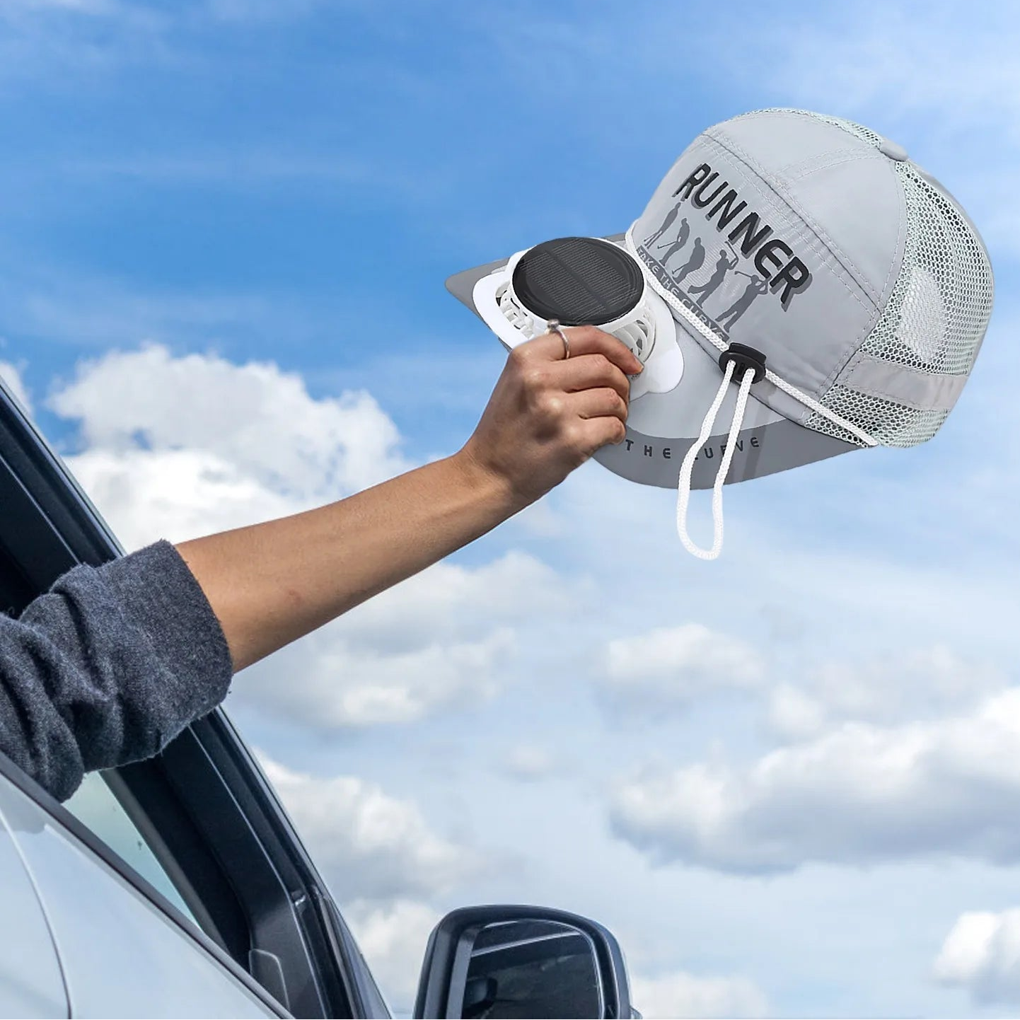 Person holding a keep cool fan cap  in the breeze outside car window, with a attached air blower against a blue sky.