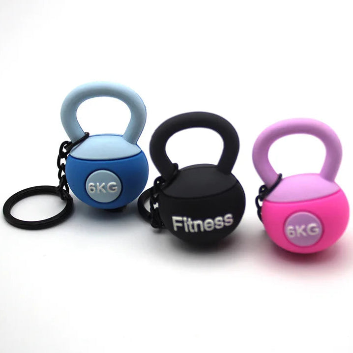 Three small kettlebell-shaped weight keychains in blue, black, and pink on a white background.