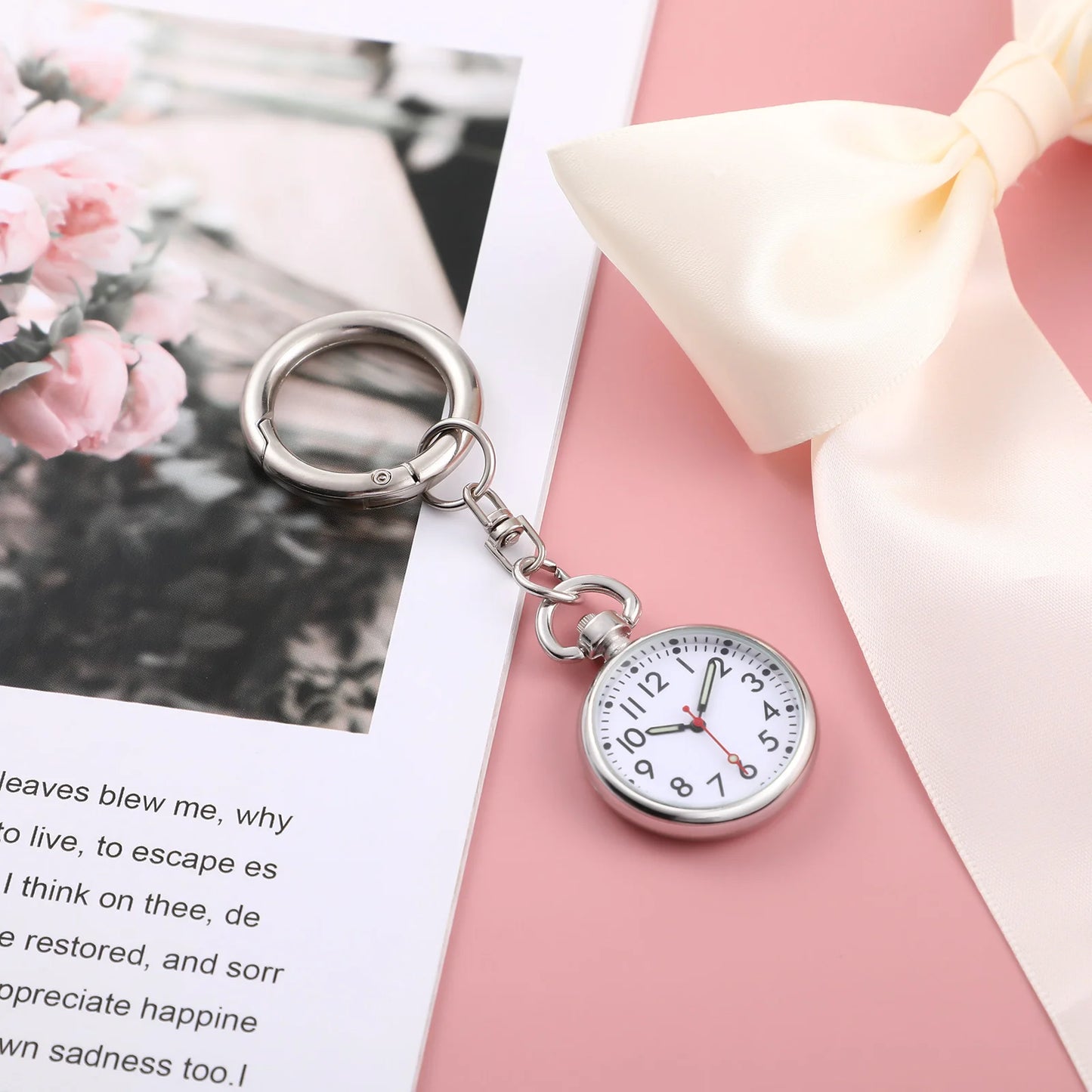 Silver pocket watch keychain on a pink surface with a blurred floral background