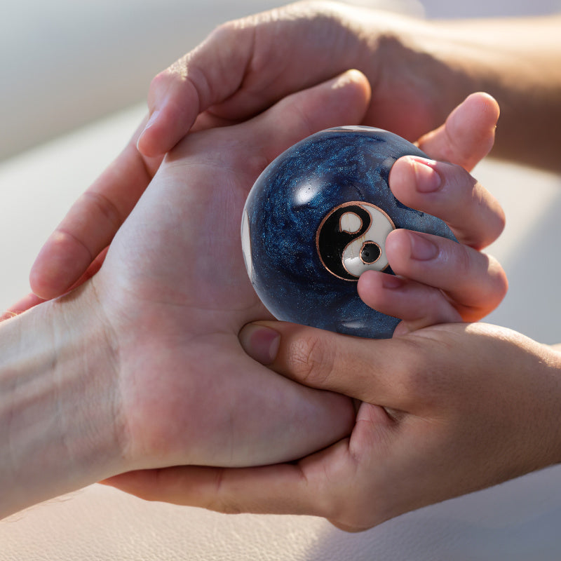A close-up of a person's hands holding a small, glossy blue marble stress ball with a black, white, and gold yin-yang symbol on the front.
