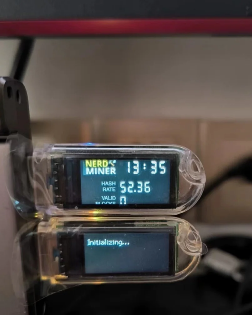 Digital display of dual plugged-in and ready mining showing 'NERD MINER' with hash rate and other metrics, placed on a reflective surface.