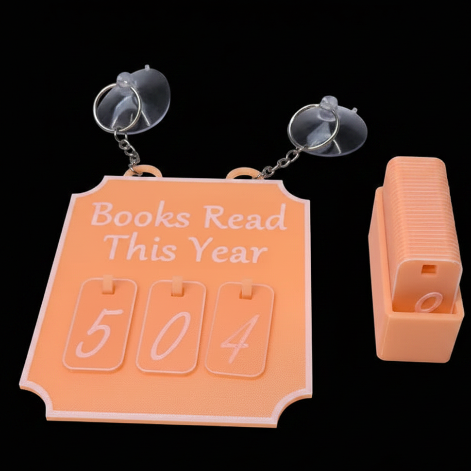 An orange wall-mounted calendar book with a suction cup on the left and a separate orange book cover or accessory on the right, shown on a black background. The main calendar reads 'Books Read This Year' with number cards featuring digits for marking.
