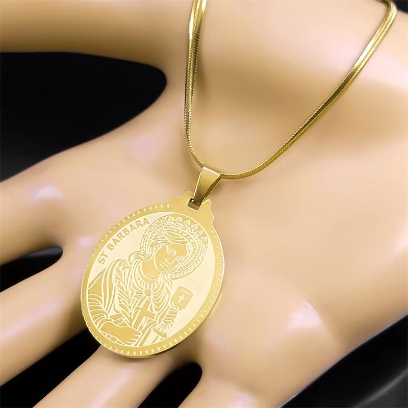 A gold-colored Saint Barbara pendant necklace with a flat chain, displayed in palm of mannequin hand over a dark background.