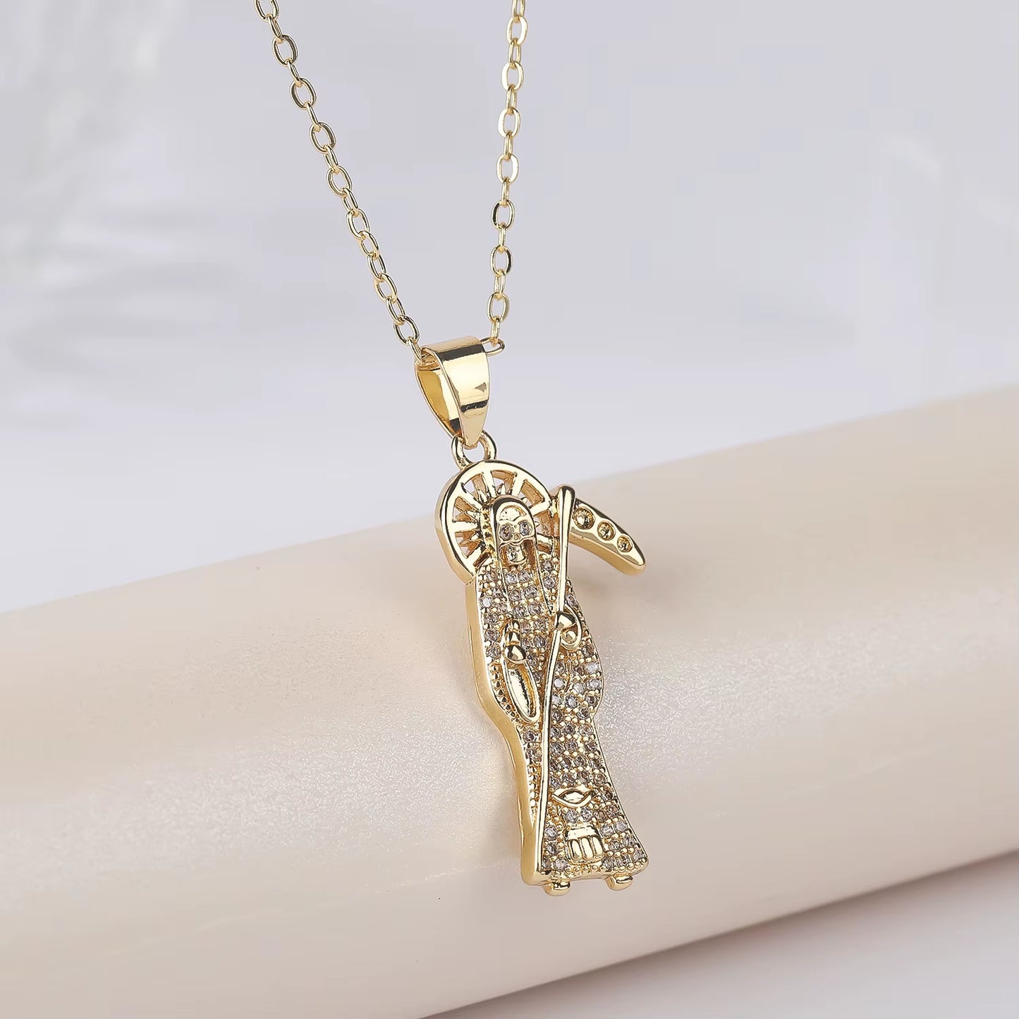 A gold-plated copper pendant in the shape of Santa Muerte holding a scythe, with cubic zirconia accents, on an adjustable chain.