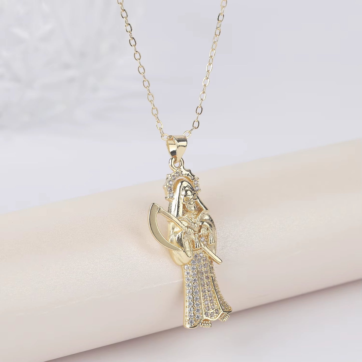 A gold-plated copper pendant in the shape of Santa Muerte holding a scythe, with cubic zirconia accents, on an adjustable chain.
