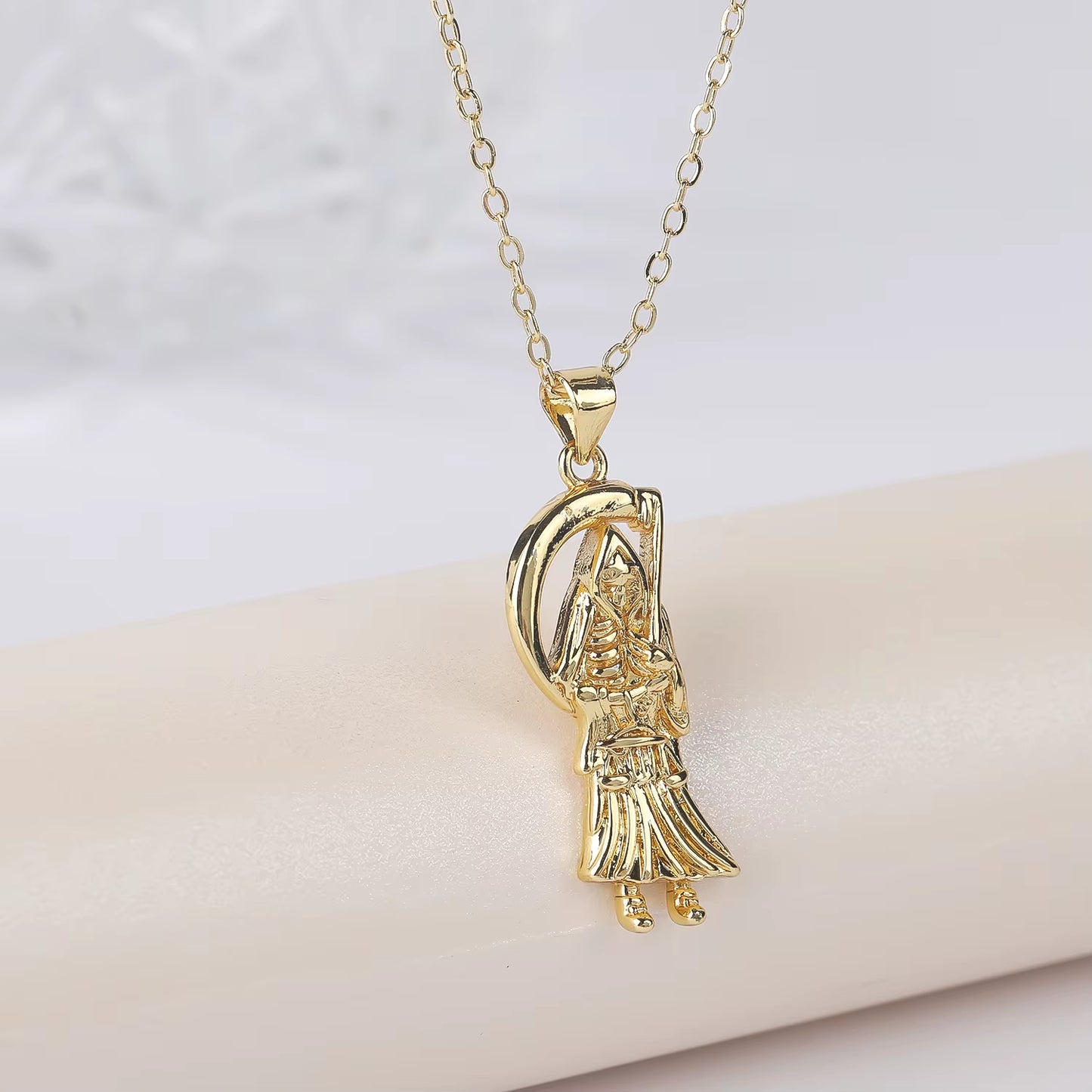 A gold-plated copper pendant in the shape of Santa Muerte holding a scythe, with cubic zirconia accents, on an adjustable chain.