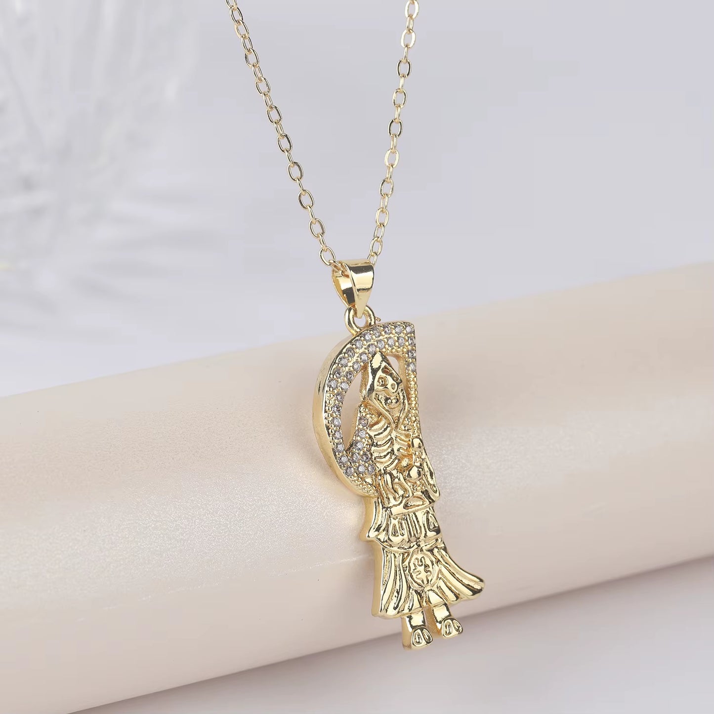 A gold-plated copper pendant in the shape of Santa Muerte holding a scythe, with cubic zirconia accents, on an adjustable chain.