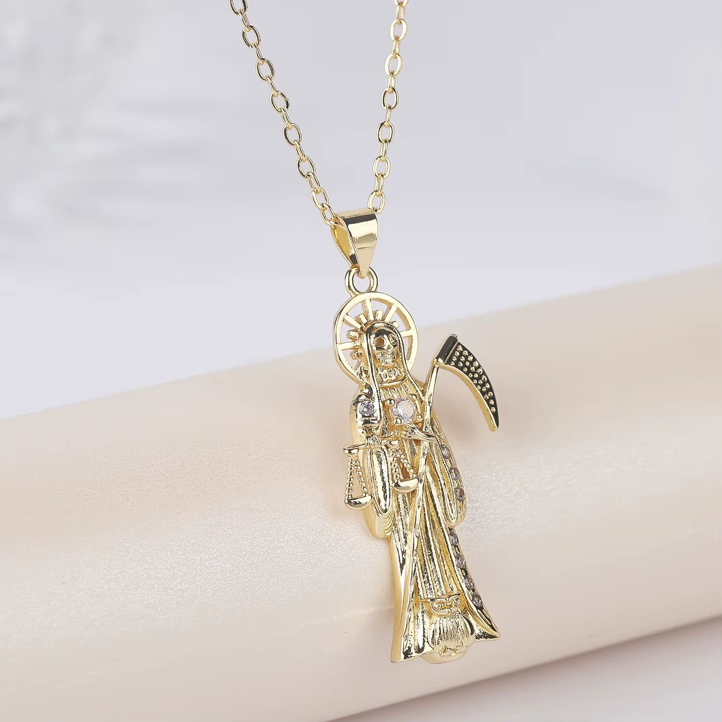A gold-color plated copper pendant in the shape of Santa Muerte holding a scythe, with cubic zirconia accents, on an adjustable chain.