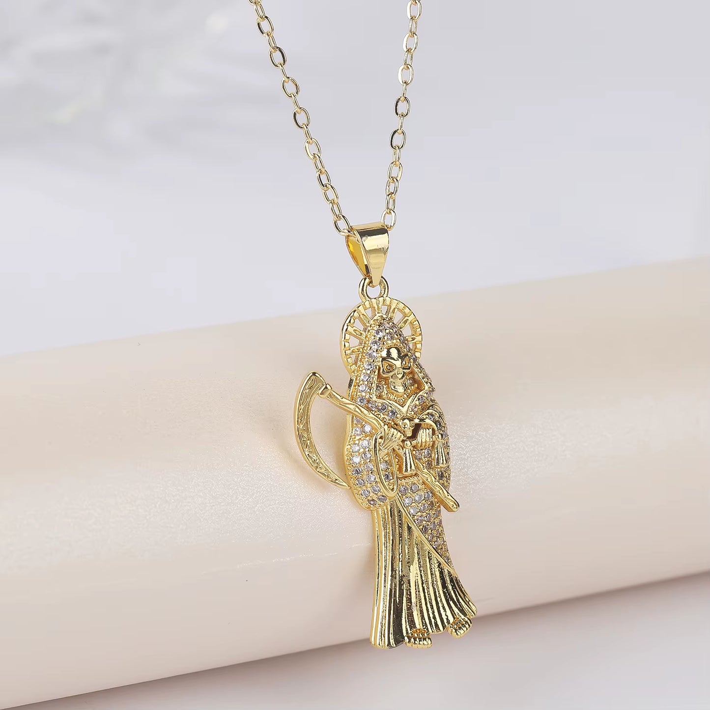 A gold-plated copper pendant in the shape of Santa Muerte holding a scythe, with cubic zirconia accents, on an adjustable chain.