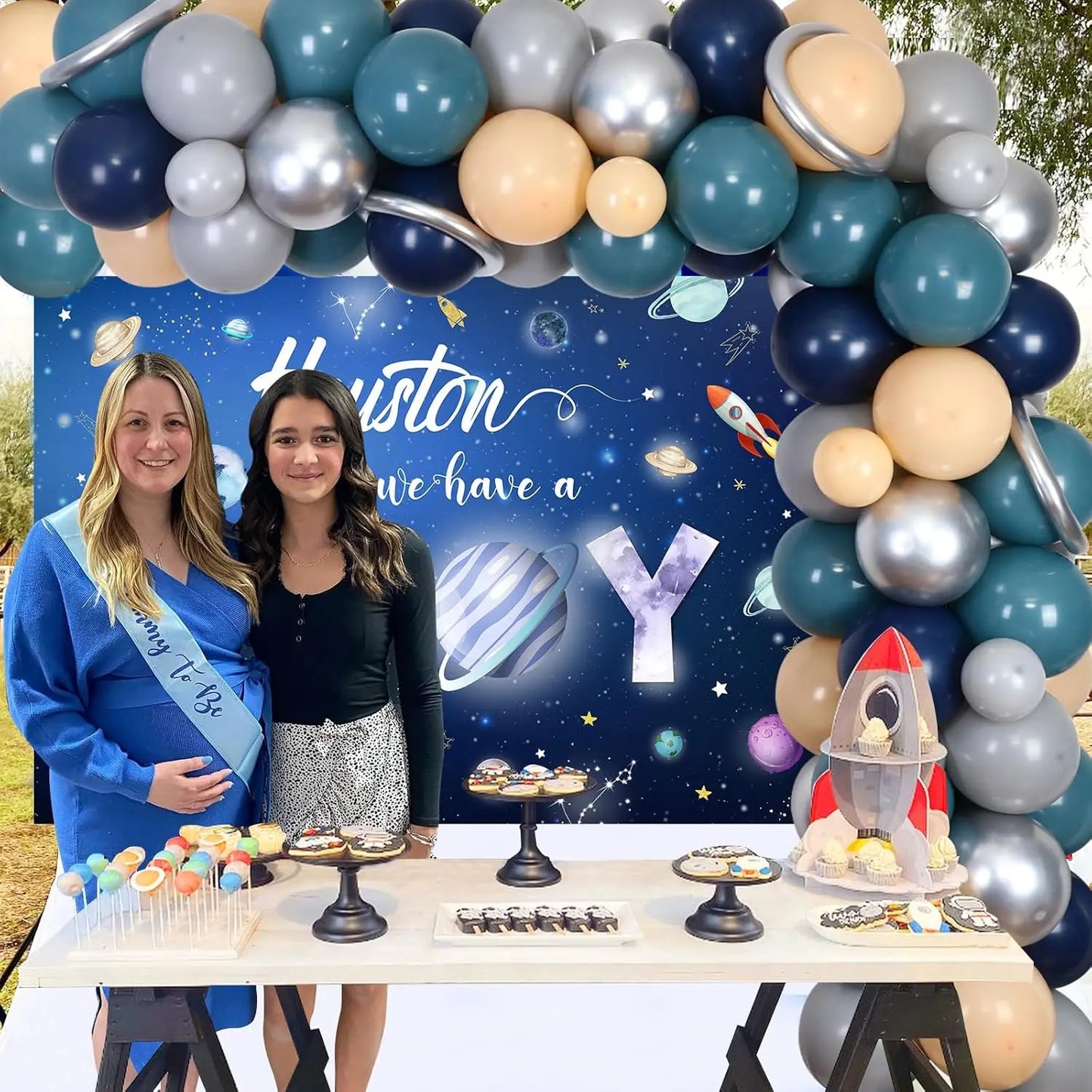Two women standing behind a table with space-themed decorations and balloons. Banner reads "Houston, We Have a Boy" with rockets and space travel elements and cosmic themed party supplies.
