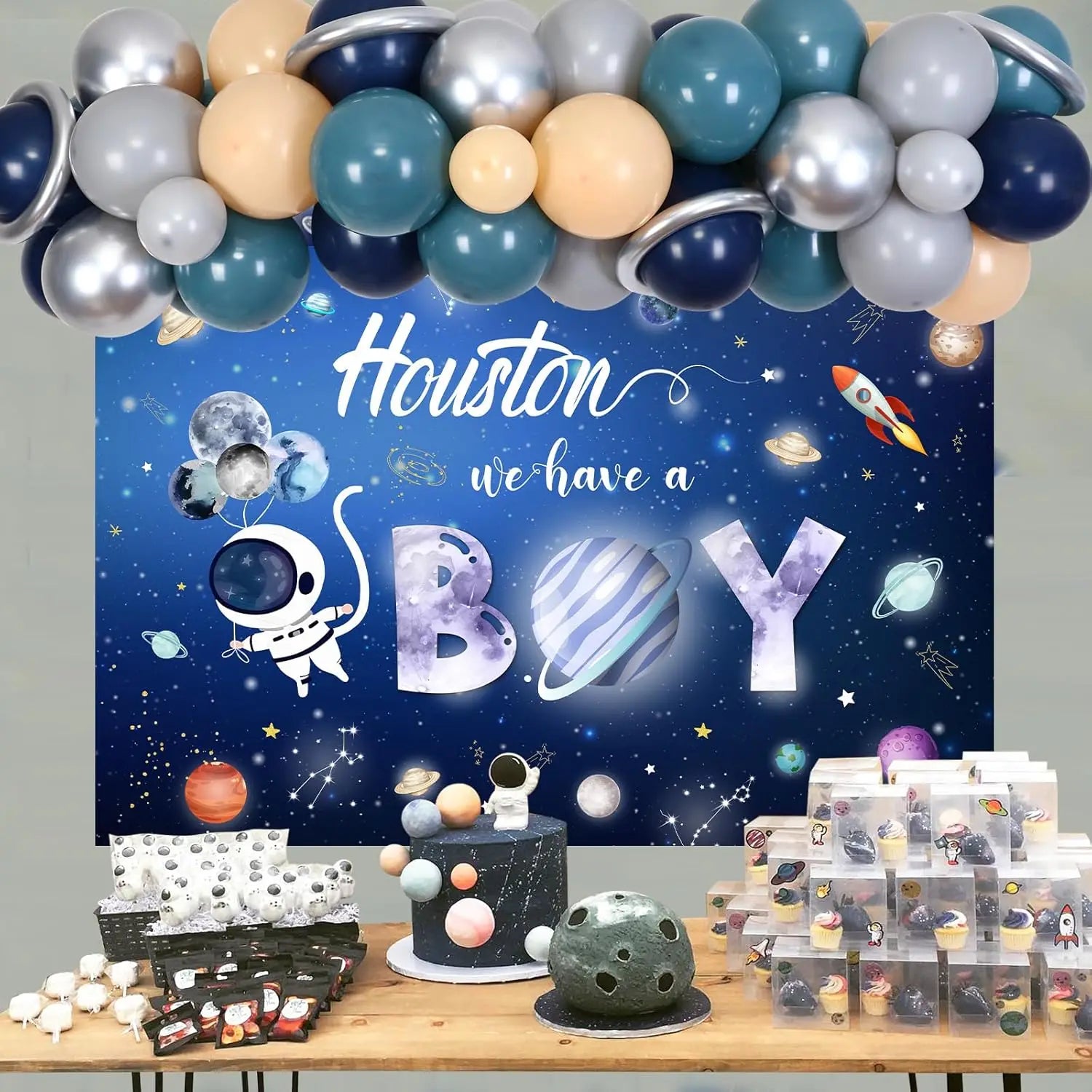 Space-themed baby shower setup with balloons, cake, and decorations against a 'Houston we have a boy' backdrop.
