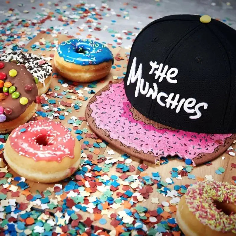 Black cap with 'the Munchies' text on a donut-shaped base with colorful donuts around, on a confetti-covered surface.